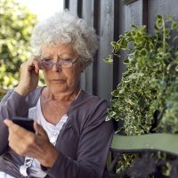 Senior lady sitting on a bench with her reading glasses in her backyard reading text message on her mobile phone